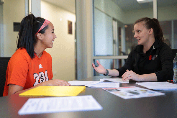 A student works with a Career Center Counselor to improve her resume. They are both sitting at a table with papers strewn about. The female student has dark hair in a pony tail and is laughing.