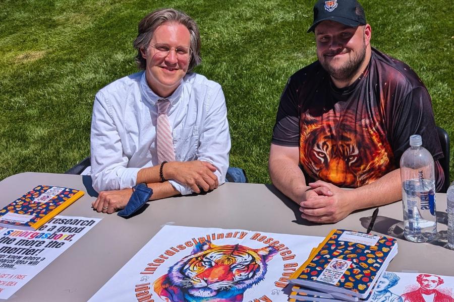 Levay and Wardell sit at a table with handouts and fliers outside on the quad