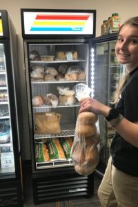 A student holds a bag of bread in front of a commercial refrigerator