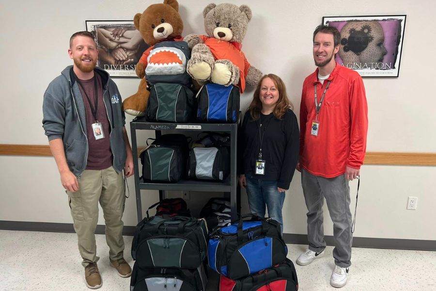 3 volunteers stand beside filled duffle bags that will be donated to local teens in need