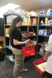 A student stands at a shelf filled with food holding a red shopping basket