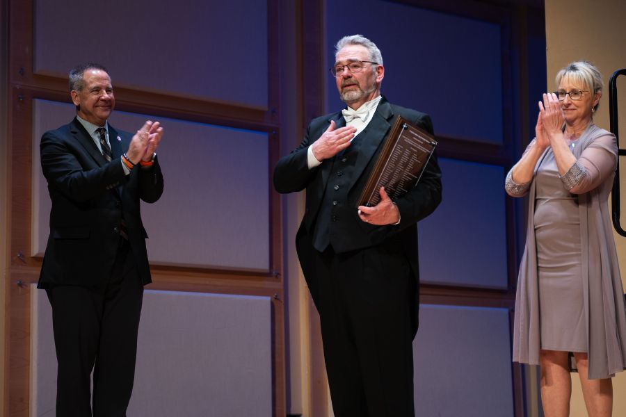 Scott Anderson stands onstage with President Wagner and Dean Turley-Ames. His hand is on his heart and he is holding a plaque.