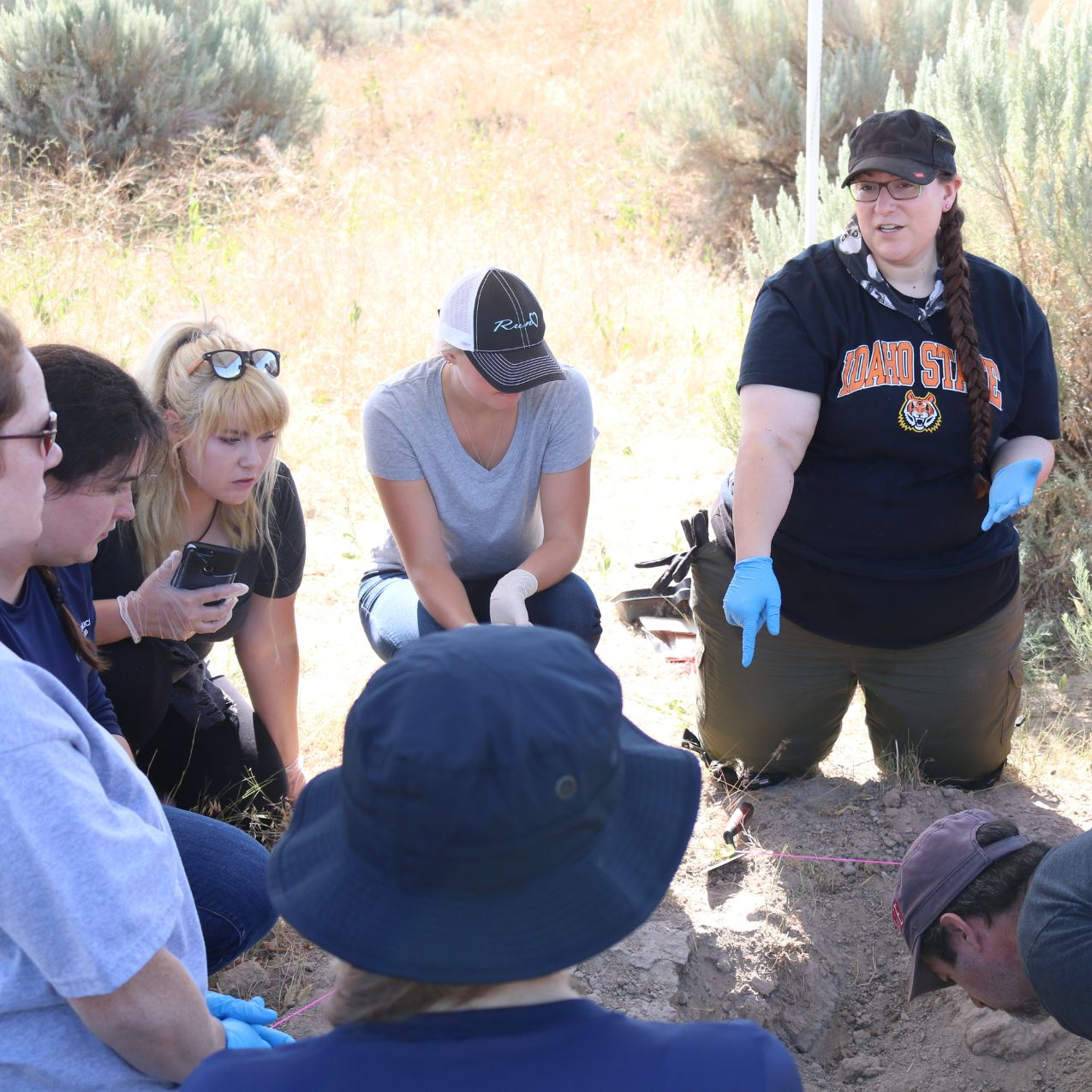Students in Samantha Blatt's class sit in a circle outside looking at bones in the ground while she points and lectures