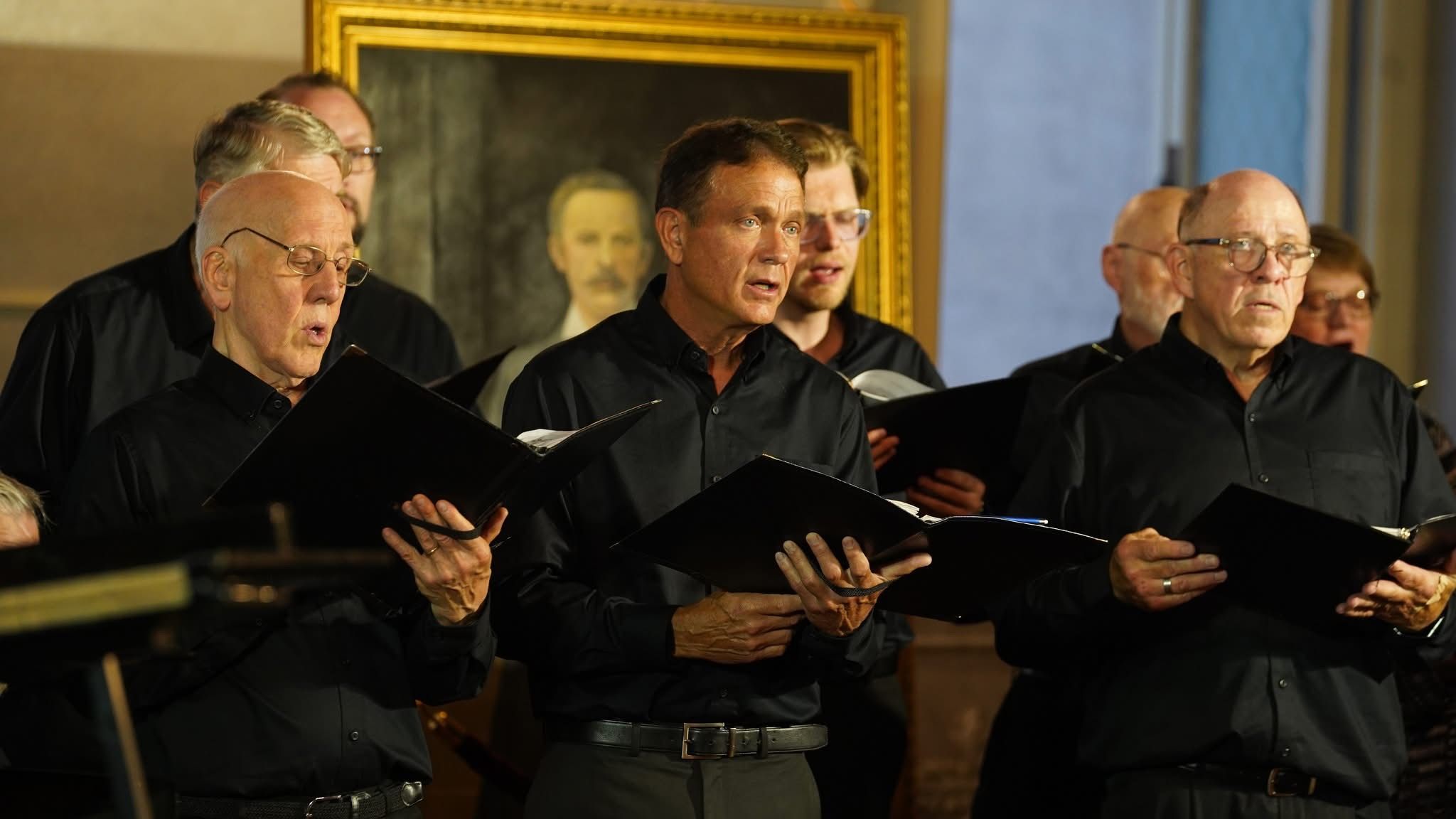 A group of male singers in black shirts performing, holding sheet music folders, in front of a large portrait.