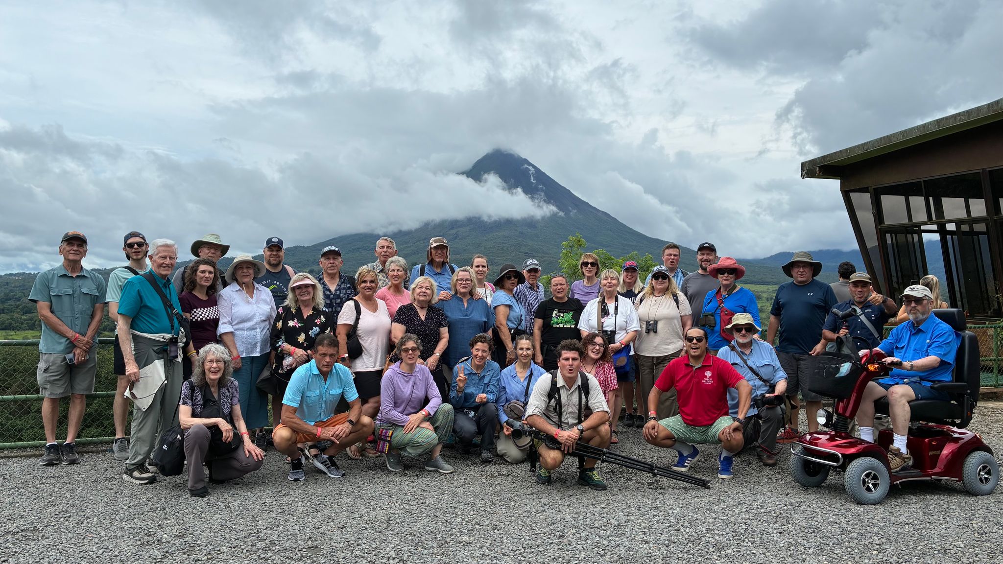 A large choir ensemble poses in front of a cloud-covered mountain, with people in casual outdoor clothing.