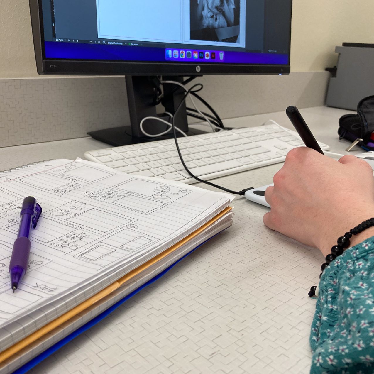 close up of a hand drawing and a notebook with a pencil and sketches. a computer is in the background