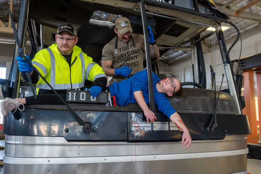 A bus is staged as an accident scene with broken windshield. An actor hangs out of the window and appears to be injured. A medical worker in a neon vest stands near the person.