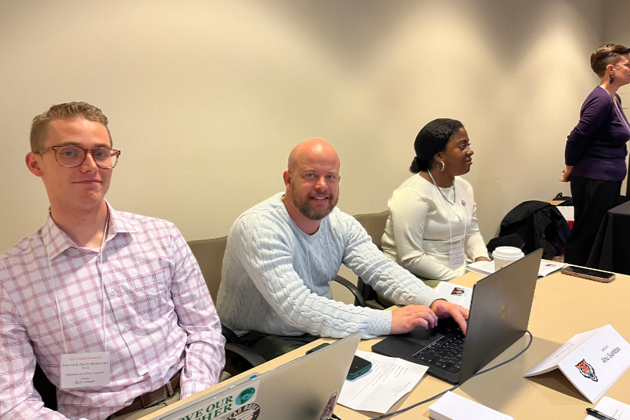 3 members of the IAC sit at a desk with laptops and smile at the camera