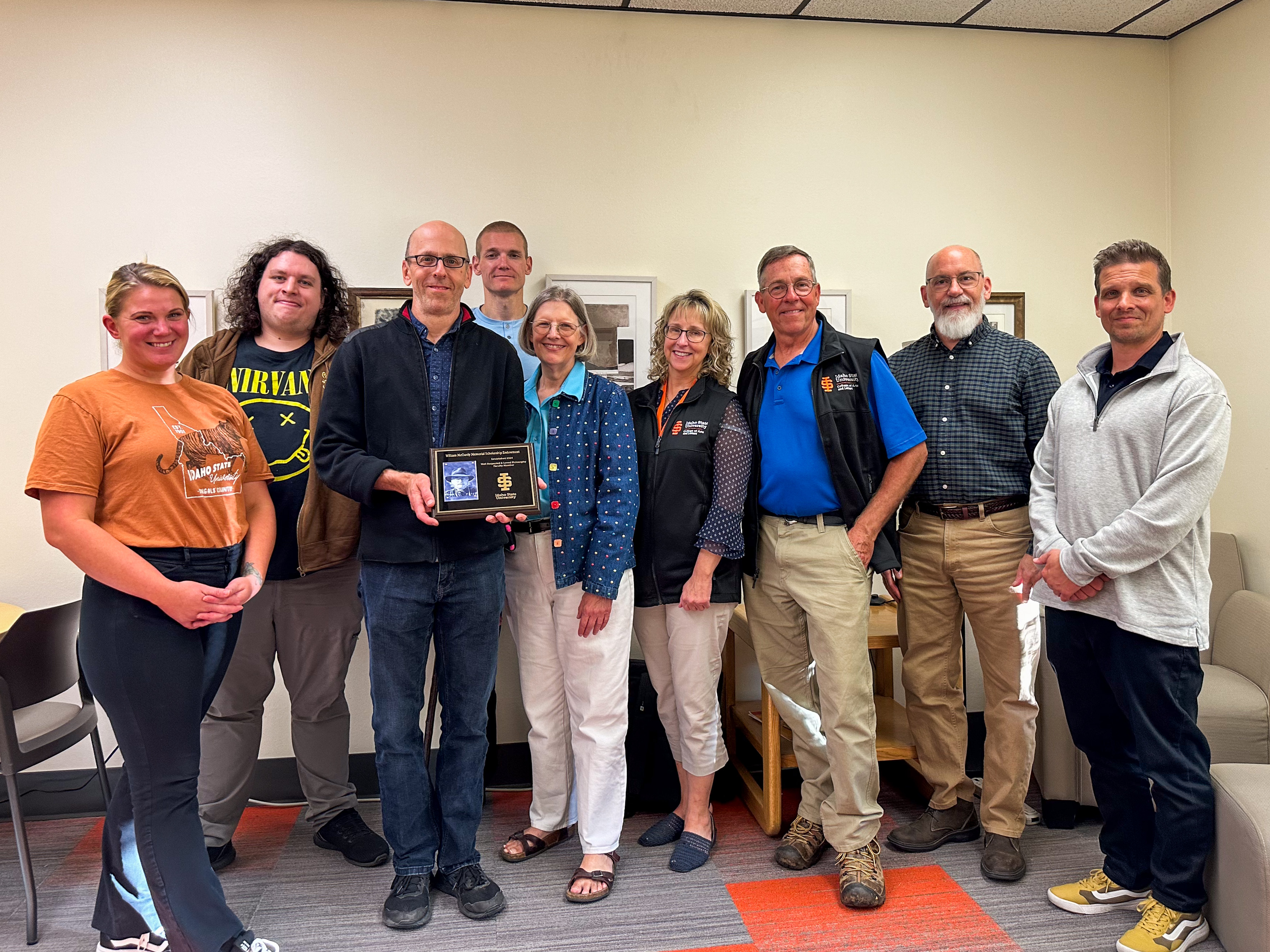 Faculty, staff, and philosophy club members stand together smiling for a group photo. Jim Skidmore holds a plaque in recognition of Will McCurdy