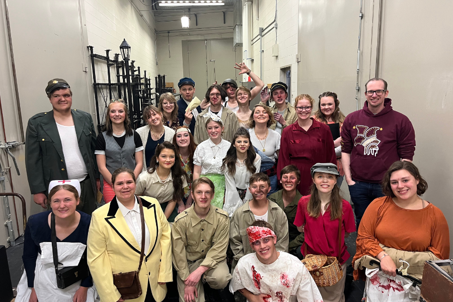 A group of students poses together backstage. They are in period makeup and costumes representing war characters. Some have stage makeup wounds.