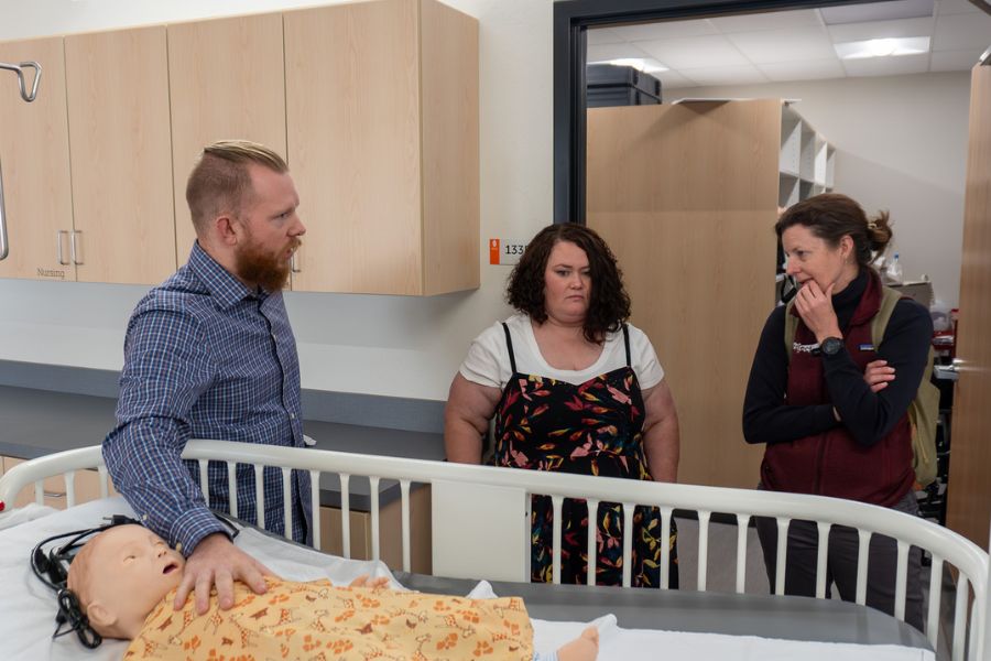 3 people stand next to a hospital bed, where a dummy lies.