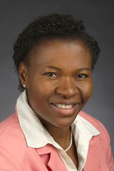 A woman with dark skin tone and short black hair. She is wearing a white collared shirt and a pink jacket