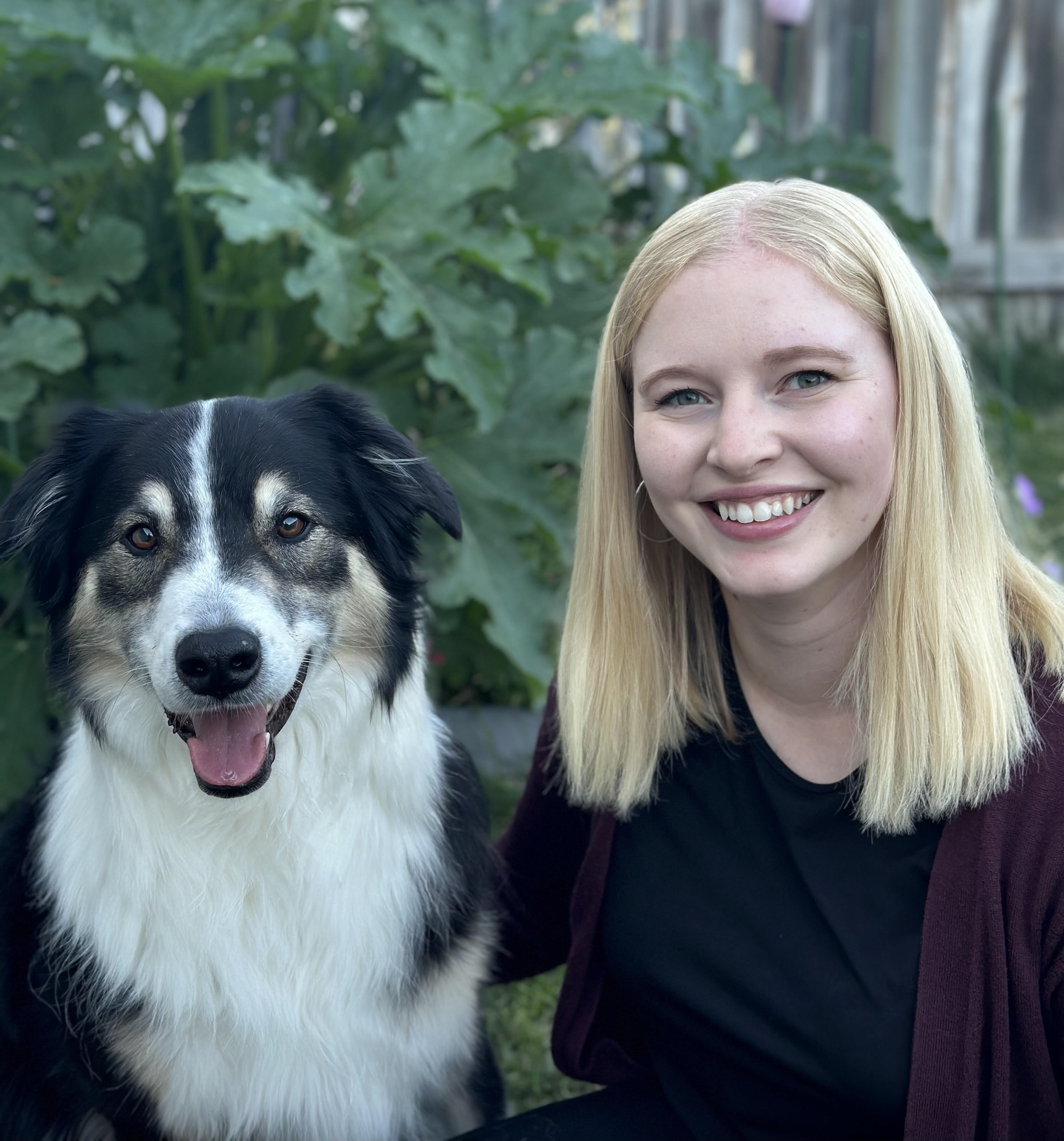 A woman with blonde, shoulder length hair; sitting with a black, white, and brown dog. The woman is smiling and the dog sits to the left of the woman with it's tongue sticking out.