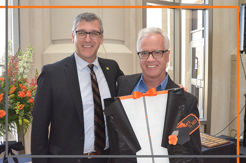 Dr. Mark Roberts holds a tuxedo shaped plaque with Dr. Rex Force in the Stephens Performing Arts Center rotunda