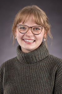 A professional portrait of Bailey Brockett wearing a green knit sweater and glasses.