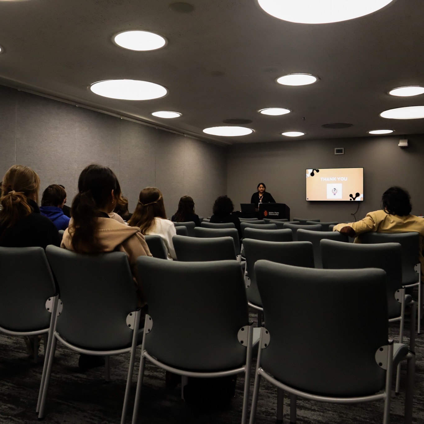 A room with people sitting in chairs while a student presents at the front from a podium