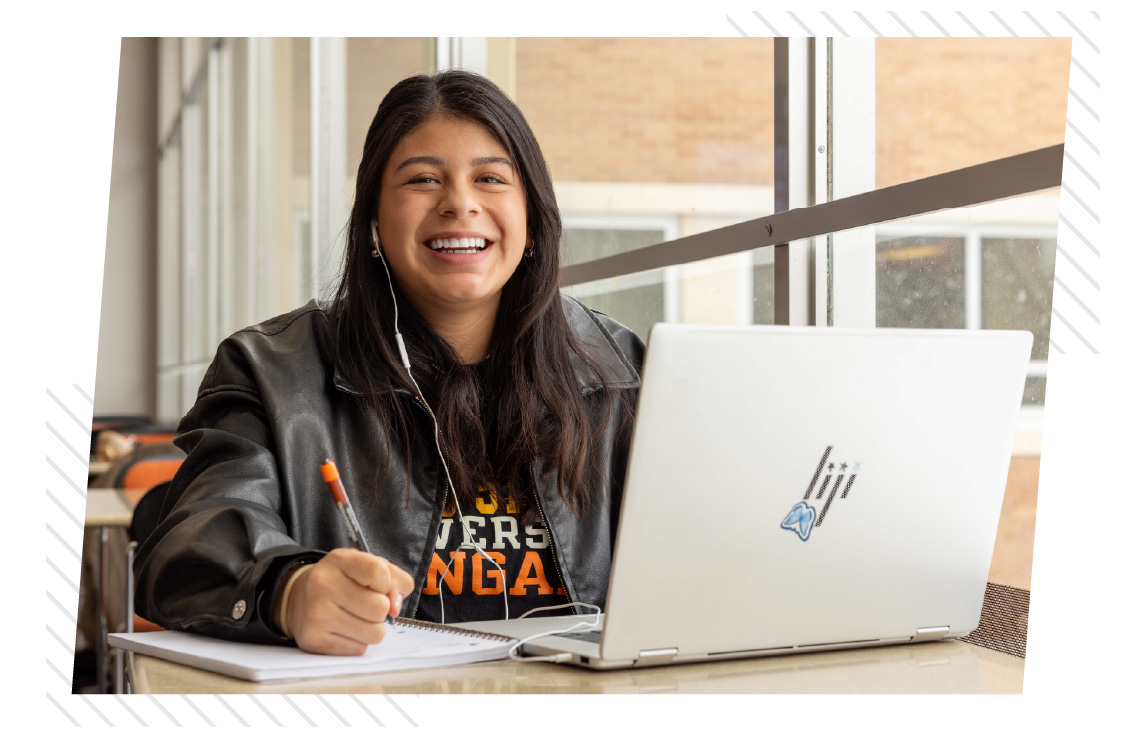 A smiling young woman sits at a table using a laptop and writing in a notebook while wearing earbuds.
