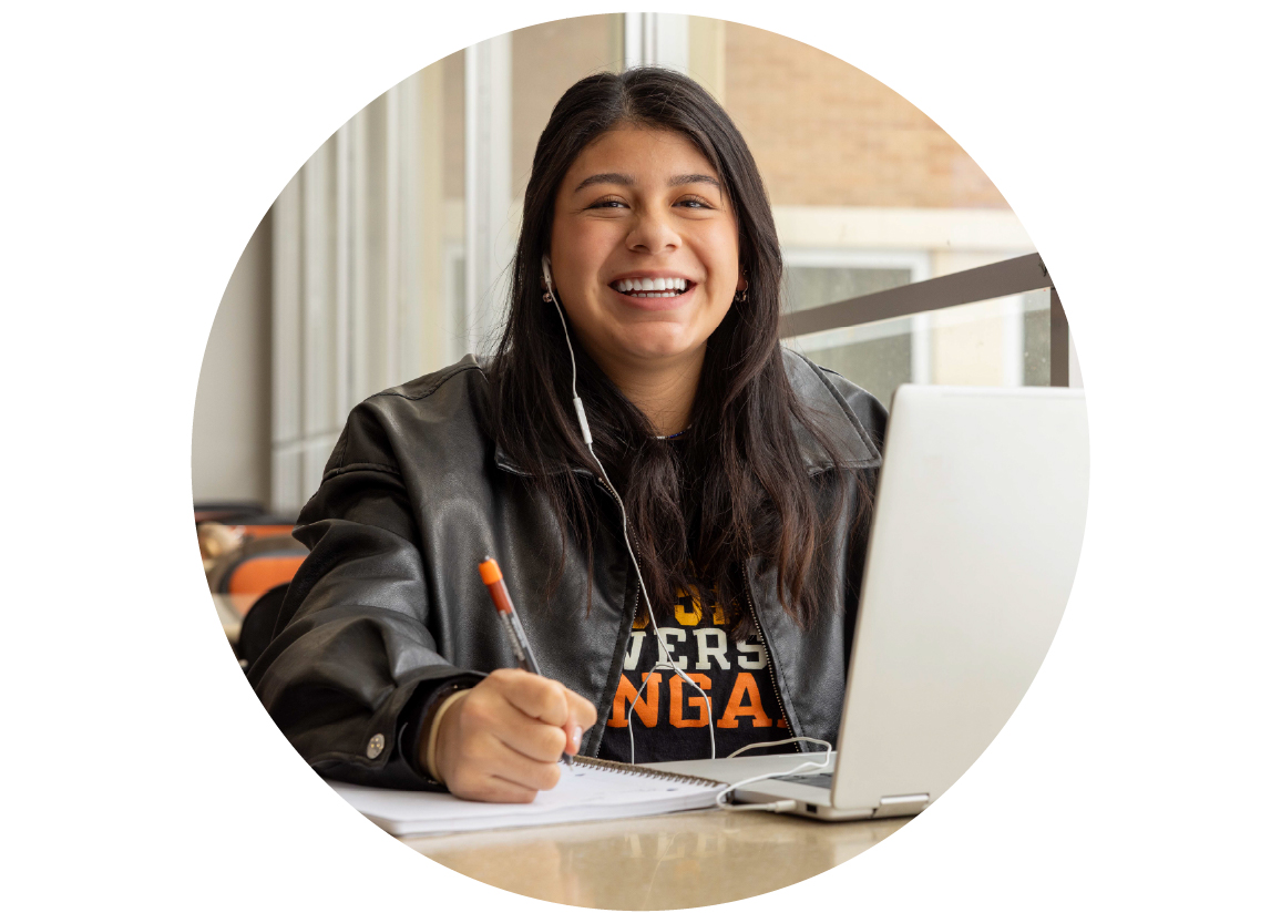 Smiling female college student sits at a table using a laptop and writing in a notebook inside a campus building.