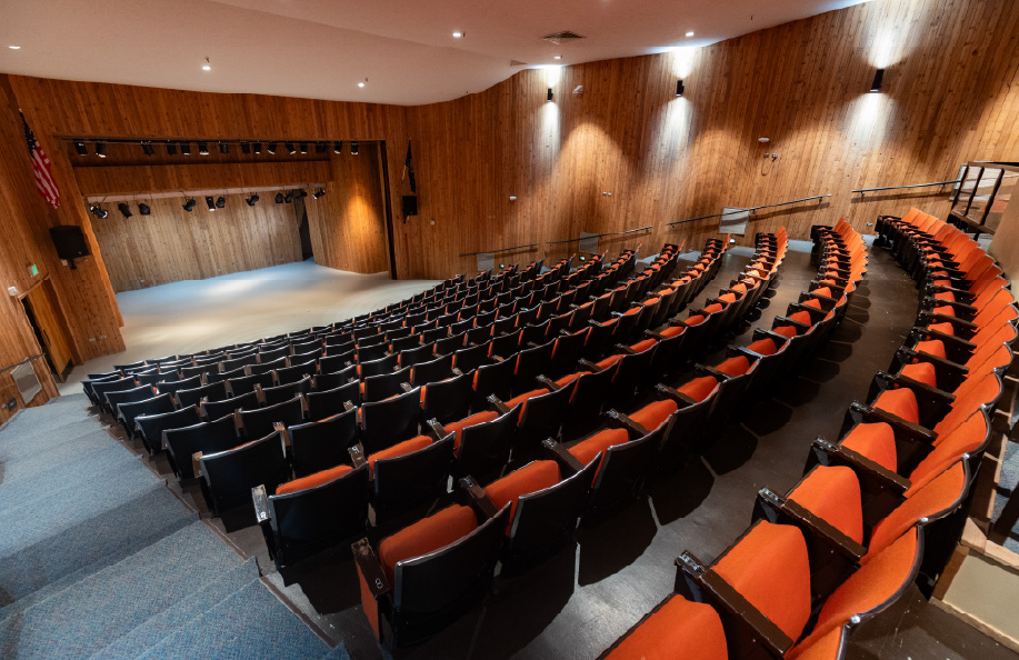 Wide interior view of a tiered auditorium with curved rows of black chairs with orange seats, facing a wood-paneled stage with overhead stage lighting; wall-mounted lights and handrails line the aisles, creating a professional venue for lectures, performances, and presentations.