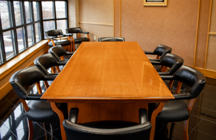 Rectangular wooden conference table surrounded by black leather chairs in a small meeting room with large windows along one wall and framed artwork on a tan wall.
