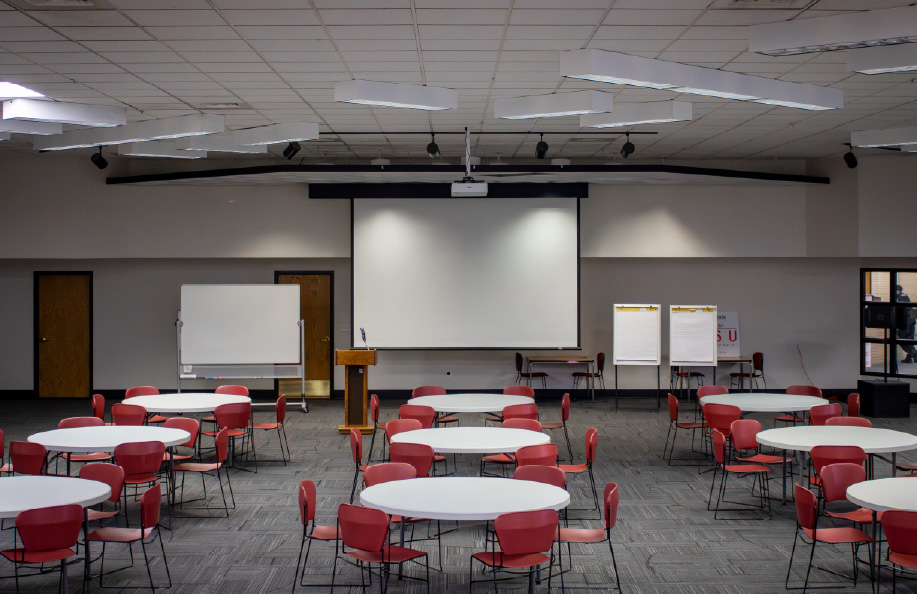 Large multipurpose room with round tables and red chairs arranged banquet-style, facing a projection screen, podium, whiteboard, and flip charts at the front of the room.
