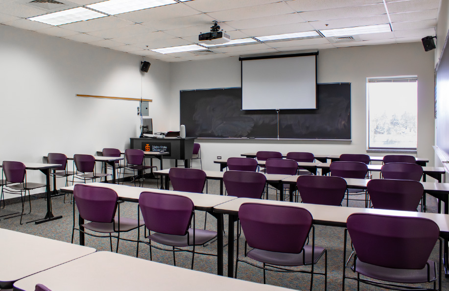 Classroom with rows of desks and purple chairs facing a chalkboard and pull-down projection screen, with an instructor podium and computer at the front.
