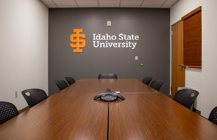 Small conference room with a wooden meeting table and black chairs, featuring an Idaho State University logo on a gray accent wall and a conference phone centered on the table.