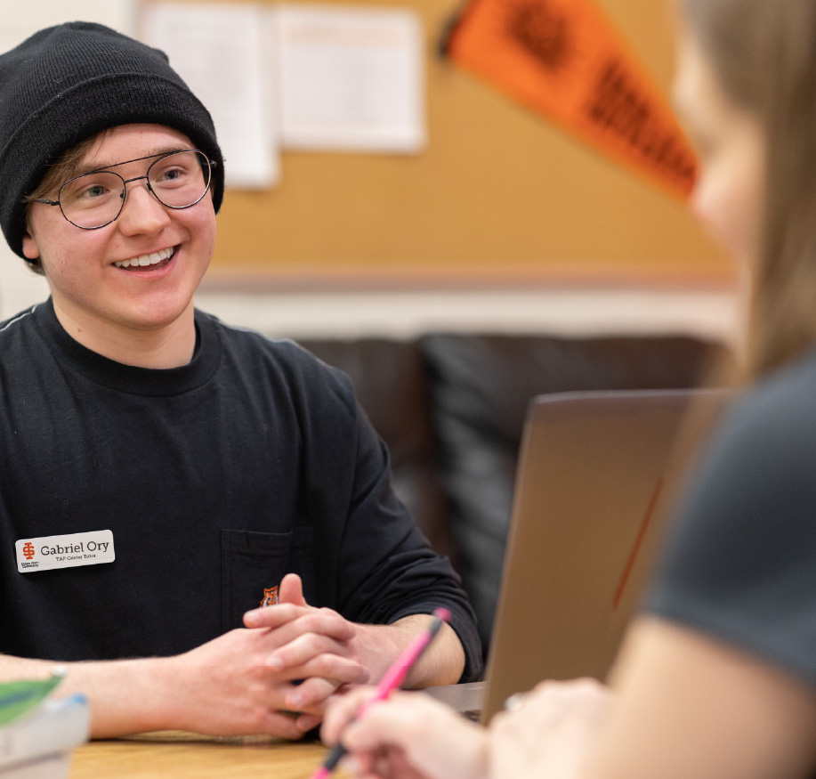 Student tutor smiling while meeting with another student at a table with a laptop during a tutoring session.