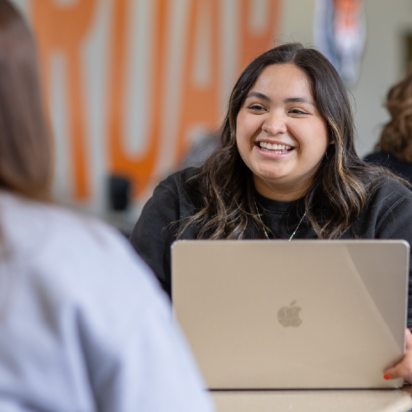 Student smiling while using a laptop and talking with another person across a table in a campus setting.