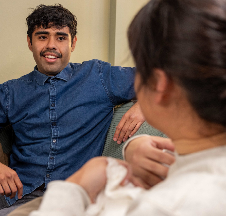 A college student sits on a couch and speaks with another person in a relaxed indoor setting. The two face each other in conversation, creating a supportive and comfortable campus environment.