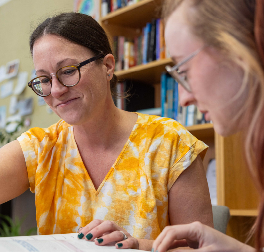 Two individuals sit together at a table reviewing paperwork in an office setting. Bookshelves line the background as they focus on the document, creating a supportive academic advising or tutoring environment.