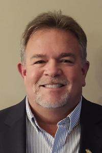 A medium skin toned man with brown and grey hair wearing a black suit jacket and and black and white striped button down shirt