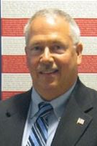 A man with medium skin tone, white and gray hair and a grey mustache. He is wearing a blue shirt, striped tie, and a black suit jacket