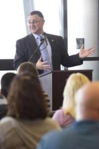 A light skin toned man standing at a podium giving a speech. He is wearing a black suit jacket, white shirt, and purple tie