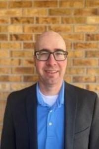 A light skin toned man who is bald, he is wearing black glasses, a blue dress shirt, and a grey suit jacket