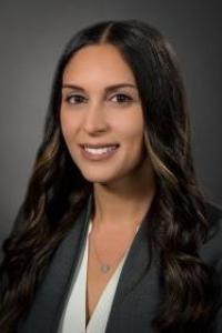 A medium-dark skin toned woman with brown hair with blond highlights. She is wearing a white blouse and grey suit jacket. 