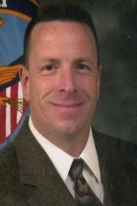 A man with medium skin tone and brown hair. He is wearing a white shirt, black tie, and grey suit jacket