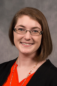 A woman with light skin tone and brown hair. She is wearing glasses, a red shirt, a necklace, and a black cardigan.