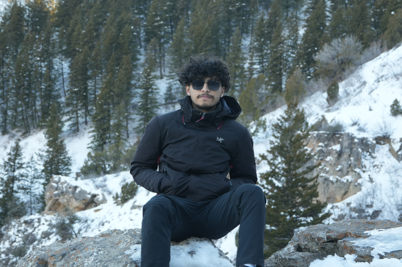 A medium shot of a young man with curly dark hair, a mustache, and sunglasses sitting on a rocky outcrop. He is wearing a black hooded technical jacket and black pants. The background is a steep, snowy mountainside filled with tall evergreen trees under bright, cold daylight.