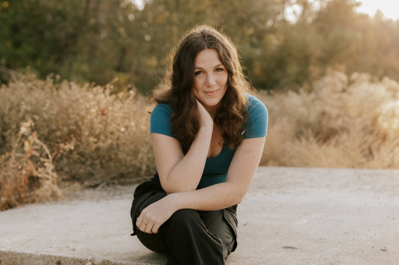 A young woman with shoulder-length, wavy dark hair and freckles poses for a portrait outdoors. She is wearing a teal t-shirt and black pants, sitting on a concrete ledge with her chin resting lightly on her hand. The background features sun-drenched, tall dry grass and soft-focus trees, captured during the 