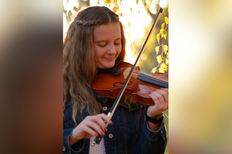 A young woman with long, wavy brown hair styled with a small crown braid plays the violin. She has a serene expression and looks down at her instrument as she plays. She is wearing a dark denim jacket over a white lace top. The background is a soft-focus autumn scene with golden yellow leaves glowing in the warm, late-afternoon sunlight.