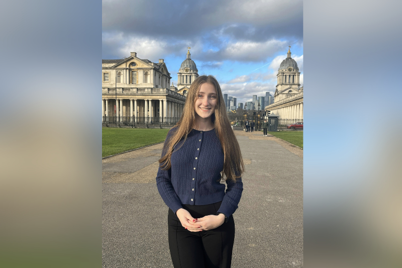 A young woman with very long, straight light brown hair smiles while standing on a paved walkway. She is wearing a navy blue cable-knit cardigan buttoned up over black trousers. In the background are the grand, classical stone buildings of the Old Royal Naval College in Greenwich, featuring prominent domes and colonnades, with a modern city skyline visible in the distance under a partly cloudy sky