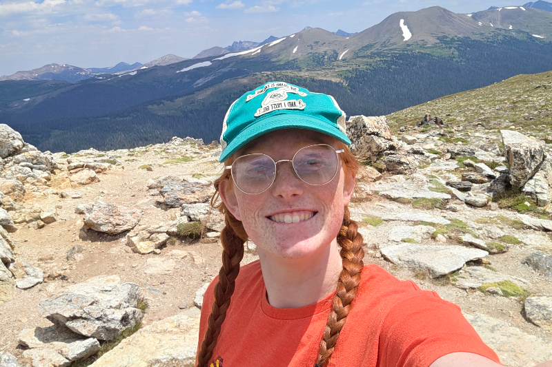 A young woman with red hair styled in two long braids smiles for a selfie on a rocky mountain summit. She is wearing an orange t-shirt, round wire-rimmed glasses, and a teal and tan trucker hat. The background features a sweeping panoramic view of a high-altitude mountain range with patches of snow on the peaks under a clear blue sky.