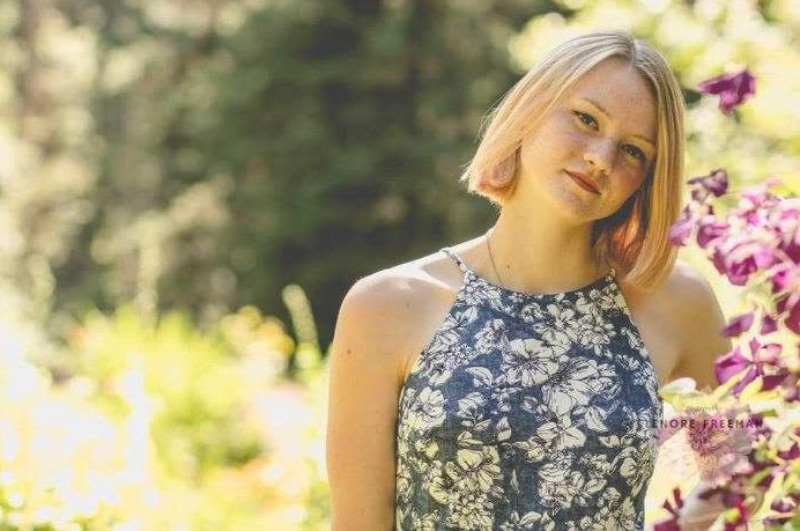 A young woman with a blonde bob hairstyle and freckles tilts her head slightly while looking at the camera with a soft expression. She is wearing a sleeveless, halter-style top with a blue and white floral pattern. She is posed outdoors in front of a soft-focus background of sunlit greenery and bright purple flowers.