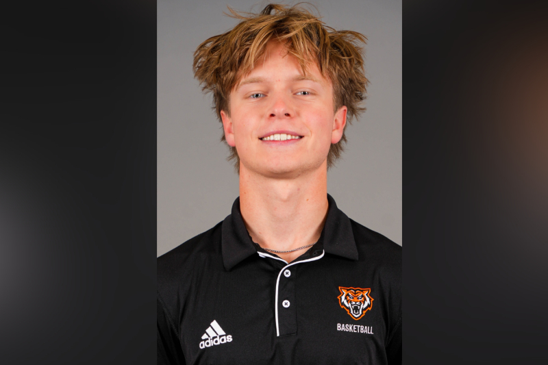 A young man with messy, light brown hair and blue eyes smiles for a headshot. He is wearing a black Adidas polo shirt featuring an orange and white tiger logo and the word 