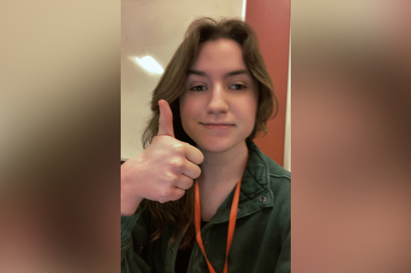 A young woman with shoulder-length brown hair gives a thumbs up directly to the camera with a subtle, confident smile. She is wearing a dark green button-up jacket and a bright orange lanyard around her neck. The background is simple, featuring a white surface on the left and a solid reddish-orange wall on the right.