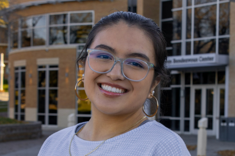 Here is the person-focused alt text for this photo:  Alt Text:  A close-up portrait of a young woman with a radiant, wide smile and dark hair pulled back. She is wearing large, clear-framed glasses, gold hoop earrings, and a delicate gold chain necklace. She is dressed in a light-colored, textured top. The background is a soft-focus view of a modern brick and glass building on a college campus.