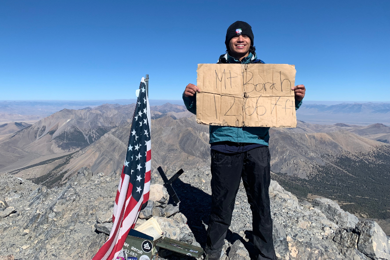 A young man stands triumphantly on a rocky mountain summit, holding a large cardboard sign that reads 