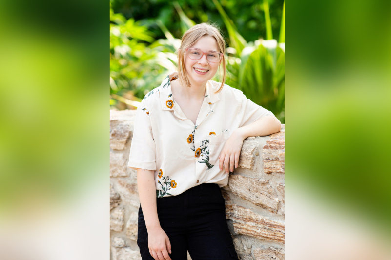 A young woman with blonde hair and pink-rimmed glasses smiles broadly in a bright, outdoor setting. She is wearing a cream-colored short-sleeved button-down shirt featuring a delicate orange floral pattern. The background is filled with lush, vibrant green tropical plants in soft focus, with sunlight filtering through the leaves.