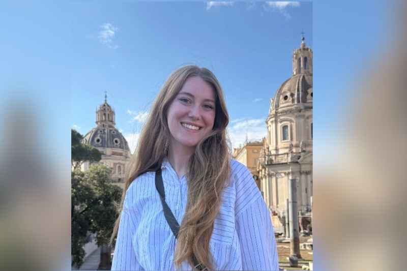 A young woman with long, wavy light brown hair smiles brightly while posing outdoors. She is wearing a white button-down shirt with thin black vertical stripes and a black crossbody bag strap. The background features grand, historic European architecture with prominent stone domes and ruins under a clear blue sky with light clouds.